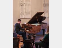 Ein Mann spielt Klavier auf einem antiken, braunen Flügel in einem kleinen Raum mit Holzboden. Im Hintergrund sind an der Wand zwei große, abstrakte Kunstwerke zu sehen, während mehrere Personen auf Stühlen sitzen und zuhören.