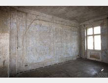 Ein leerer Raum mit einer verblassten und abgenutzten Wand in Grau- und Blautönen, einem Fenster mit Holzrahmen auf der rechten Seite und einem Betonboden. Sichtbare Rohre verlaufen entlang der Wand unterhalb des Fensters.