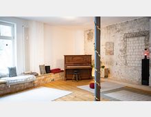 Heller Innenraum im Loft-Stil mit Holzfußboden, weiß gestrichenen Ziegelwänden und einer freigelegten, teils verputzten Backsteinwand. An der Fensterseite steht eine gepolsterte Sitzbank mit Kissen; in der Mitte ein Klavier mit Hocker sowie ein Metallstützpfeiler, daneben Teppiche und ein Lautsprecher.