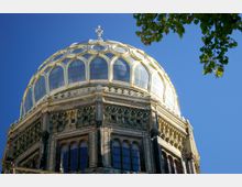 Kuppel einer historischen Synagoge mit goldenen Verzierungen und Ornamenten, darunter eine Fassade aus Backstein mit Rundbogenfenstern und dekorativen Elementen; im Hintergrund ein klarer blauer Himmel und am Rand ein Zweig mit Blättern.