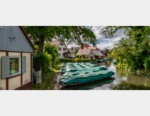 Ein kleiner Kanal mit grün abgedeckten Booten, gesäumt von Bäumen, Hecken und traditionellen Häusern mit roten Ziegeldächern. Links im Bild steht ein Fachwerkhaus, während der Kanal durch eine idyllische dörfliche Umgebung führt.