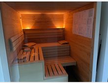 Blick in eine kleine Holzsauna mit Glasfront, umlaufenden Holzbänken in Stufenform und warmen indirekten Wandlichtern. An der rechten Wand hängt ein großer Plan, vorne steht eine niedrige Bank oder Ablage.