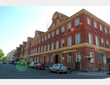 Backsteinbau einer historischen Straßenecke mit drei Etagen, auffälligem Ziergiebel und gelben Fensterrahmen. Im Erdgeschoss befindet sich eine Gaststätte mit der Aufschrift „Zum Langen Kerl“, davor sind geparkte Autos und Fahrräder sichtbar.