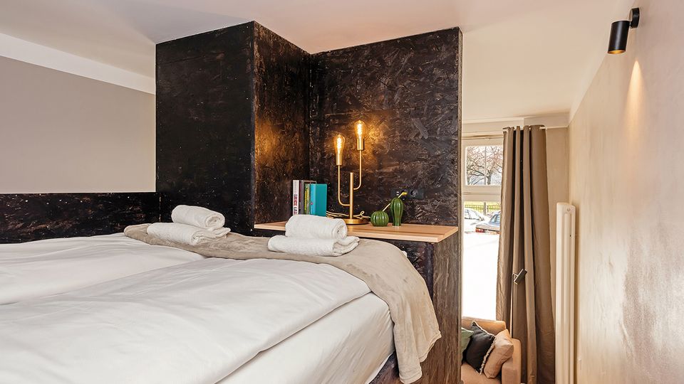 Modernes Schlafzimmer mit großem Bett und weißer Bettwäsche, integriert in eine dunkle, holzverkleidete Wandstruktur mit Ablage, Büchern und zwei Leuchten. Rechts befindet sich ein Fenster mit schweren Vorhängen sowie eine schmale Sitznische und ein Heizkörper an der Wand.