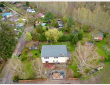 Luftaufnahme eines Wohngebiets mit einem zweistöckigen Haus mit Flachdach im Vordergrund, umgeben von einem großen Garten mit Bäumen und einem kleinen Teich in Bau. Im Hintergrund sind weitere Kleingärten, Wege und kleinere Gebäude in ländlicher Umgebung zu sehen.