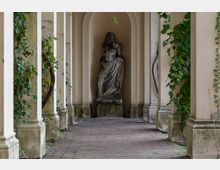 Ein überdachter Gang mit cremefarbenen Säulen und Kletterpflanzen, die teilweise die Wände bedecken. Am Ende des Gangs befindet sich eine Nische mit einer alten Steinstatue, die einen teilweise zerbrochenen Körper in einem drapierten Gewand darstellt.