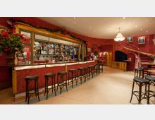 Innenraum einer eleganten Bar mit geschwungener Theke, hohen Holzstühlen und einer rot-goldenen Farbgestaltung. Im Hintergrund befinden sich ein Kronleuchter, ein Treppenaufgang mit rotem Teppich und dekorative Blumenarrangements.