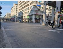 Straßenkreuzung in einer Stadt mit mehrstöckigen, modernen Gebäuden im Hintergrund, darunter Geschäfte wie „shoe!tz city“ und „Timberland“. Fußgänger und Fahrradfahrer überqueren die Straße bei einer grün markierten Ampel.