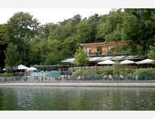 Ein Café mit Terrasse und weißen Sonnenschirmen am Ufer eines Sees, umgeben von dichtem Wald. Das Gebäude besteht aus Holz mit großen Fenstern und einem Balkon, auf dem Blumen stehen.