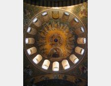 Deckengemälde einer Kuppel in einer Kirche, verziert mit goldenen Mosaiken, himmlischen Figuren und kunstvollen Ornamenten. Mehrere kleine Fenster lassen Licht herein und beleuchten die detailreiche Darstellung.