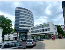 Mehrstöckiges Bürogebäude mit einer abgerundeten Fassade auf der linken Seite und einem flachen, rechteckigen Gebäude mit Geschäften und einem Café im Erdgeschoss rechts. Im Vordergrund zahlreiche parkende Autos, Fahrräder und ein Gehweg; im Hintergrund Bäume und bewölkter Himmel.