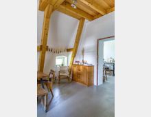 Wohnraum in einem rustikalen Stil mit hellen Wänden und einer sichtbaren Holzbalkenkonstruktion an der Decke. Der Raum verfügt über eine Holzkommode, einen Holztisch mit passenden Stühlen sowie ein kleines Fenster mit Dekorationen; im Hintergrund ist ein Durchgang zu einem weiteren Zimmer sichtbar.