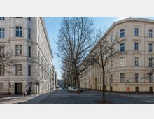 Straßenszene mit zwei gegenüberliegenden Altbauwohnhäusern in heller Fassade, getrennt durch eine baumbestandene Straße. Im Vordergrund ist eine Person mit Kinderwagen zu sehen, daneben geparkte Autos und Straßenlaternen.