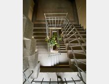 Treppenhaus in einem mehrstöckigen Gebäude mit grauen Betonstufen und Metallgeländern, im Zentrum ein Blick nach unten auf einen bepflanzten Bereich in der Mitte. Die Umgebung ist dezent beleuchtet, und der Boden im unteren Bereich besteht aus cremefarbenen Fliesen.