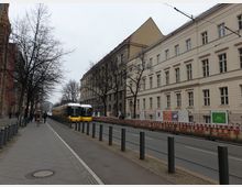 Straße mit zweigleisiger Straßenbahnlinie, auf der ein gelb-weißer Straßenbahnzug fährt. Links stehen Bäume und ein Gehweg mit Passanten, rechts sind hellbeige mehrstöckige Gebäude im klassischen Stil sowie eine Baustellenabsperrung sichtbar.
