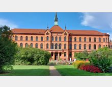 Backstein-Gebäude mit drei Stockwerken und einem Turm mit Uhr, umgeben von einem Garten mit gepflastertem Weg, Rasenflächen, Sträuchern und bunten Blumenbeeten. Der Himmel ist klar mit wenigen Wolken.