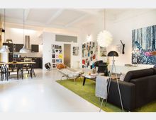 Offener Wohnraum mit modernem Design, bestehend aus einer Küche mit dunklen Schränken und einem Esstisch links sowie einem Wohnzimmerbereich rechts mit schwarzem Sofa, Regalen und Kunstwerken an der Wand. Der Raum ist hell, hat weiße Wände und Decken sowie einen grünen Teppich im Wohnbereich.