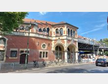 Backstein-Gebäude im neoklassizistischen Stil mit Rundbogenfenstern und dekorativen Steinelementen. Rechts daneben führt eine erhöhte U-Bahn-Trasse entlang, mit einem Zugang zur U-Bahn-Station "Schlesisches Tor". Fahrräder stehen entlang des Gehwegs, und einige Autos sind auf der Straße zu sehen.
