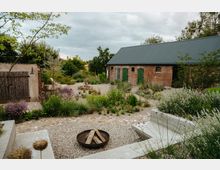 Ein rustikaler Innenhof mit Kiesboden und Steinwegen, umgeben von üppiger Bepflanzung mit Gräsern und Blumen. Im Hintergrund stehen ein Backsteingebäude mit grünen Holztüren und ein kleiner Schuppen mit Holzverkleidung, daneben eine offene Feuerstelle.