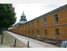 Gelb gestrichenes historisches Gebäude mit langen Fassaden und mehreren Fenstern, flankiert von einem gepflasterten Gehweg. Im Hintergrund ein Turm mit grünem Kupferdach, Uhr und einer Adlerstatue an der Spitze.