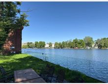 Uferbereich mit Blick auf einen Fluss, umgeben von einer Glasbrüstung. Links steht eine rote Ziegelwand neben einer Wiese, auf der Stühle sichtbar sind. Im Vordergrund befindet sich ein Tisch mit Stühlen, im Hintergrund sind Bäume, Gebäude und ein Bootshaus am gegenüberliegenden Ufer zu sehen.