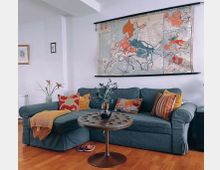 Wohnzimmer mit einer dunkelgrauen Couch, die mit bunten Kissen und einer gelben Decke dekoriert ist. Über der Couch hängt eine große historische Europakarte, der Holzboden ist sichtbar, und vor der Couch steht ein kleiner runder Metalltisch mit einer Vase mit Blumen darauf.