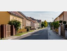 Eine ruhige Wohnstraße mit Einfamilienhäusern auf beiden Seiten, die meist verputzt und in gedeckten Farben gestrichen sind. Die Straße ist asphaltiert, von schmalen Gehwegen mit Pflastersteinen gesäumt und wird von Vorgärten mit Zäunen und Bäumen begrenzt.