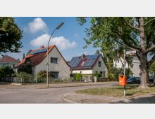 Straßenszene mit Einfamilienhäusern, die rote Ziegeldächer und Solarpaneele auf dem Dach haben. Im Vordergrund eine Laterne, ein Baum und ein Mülleimer entlang des Gehwegs; Straßenschild mit der Aufschrift „Birnhornweg“ sichtbar.