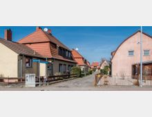 Straßenszene einer Wohnsiedlung mit Einfamilienhäusern, die überwiegend beige Fassaden und rote Walmdächer haben. Im Vordergrund stehen Straßenschilder und Zäune, während der Weg zwischen den Häusern in Richtung weitere Gebäude führt.