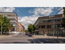 Straßenszene mit Kreuzung, flankiert von modernen Bürogebäuden mit großen Fenstern auf der rechten Seite und Bäumen sowie weiteren Gebäuden links. Im Vordergrund Ampeln und Straßenschilder, dahinter parkende Autos und eine von beiden Seiten gesäumte Straße.