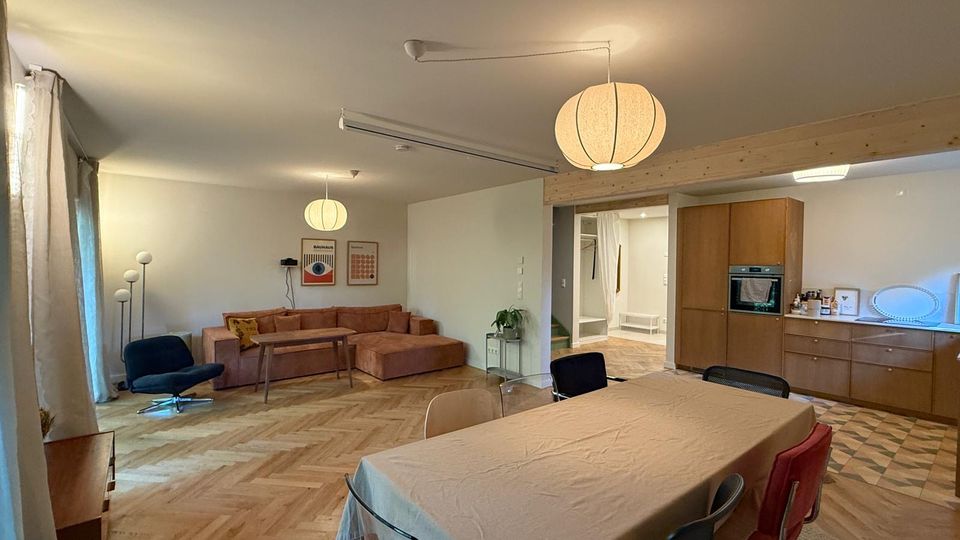 Offener Wohnbereich mit Holzparkettboden, einer großen Eckcouch in Braun und einem Esstisch mit mehreren Stühlen. Im Hintergrund befinden sich eine Küche mit Holzschränken und ein Eingangsbereich mit minimalistischer Einrichtung.