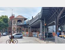 Straßenkreuzung mit historischem U-Bahn-Eingangsgebäude aus rotem Backstein und hellen Steinelementen, daneben eine Hochbahntrasse aus Metall. Im Vordergrund sind Fußgänger, ein Radfahrer und Autos zu sehen, während die Umgebung mit Graffiti und Werbetafeln geprägt ist.