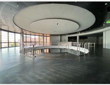 Weitläufiger, moderner Innenraum mit schwarzem Boden und einer großen runden Deckenstruktur. In der Mitte befindet sich eine metallene Wendeltreppe mit Geländer, umgeben von Fenstern, die Tageslicht hereinlassen.