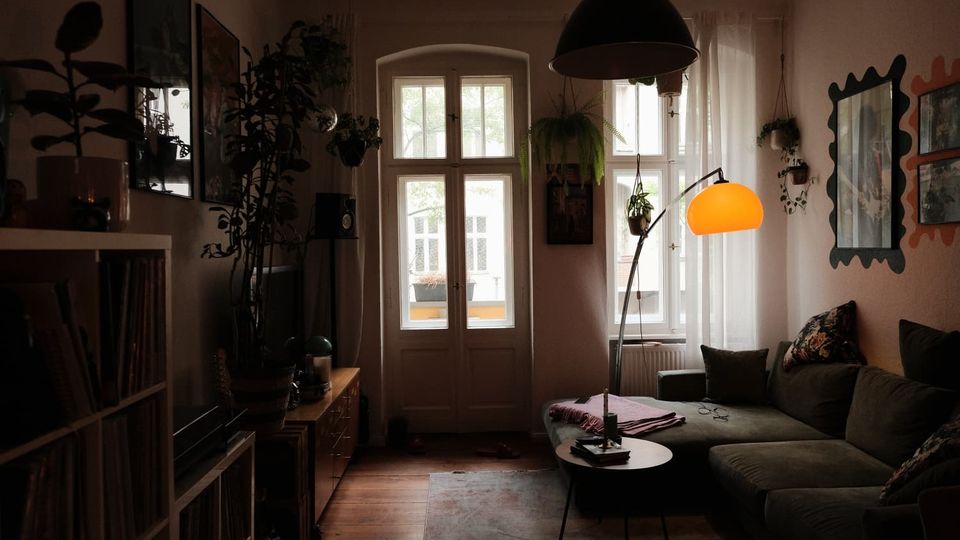 Gemütliches Wohnzimmer mit hohen Fenstern und einer Glastür zum Balkon, davor ein Sofa mit Couchtisch und Stehlampe. Pflanzen hängen an der Decke und an den Wänden; links stehen Regale und ein Lowboard, rechts Wandspiegel und Bilder.
