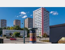 Mehrere Hochhäuser mit rot-grauen Fassaden stehen im Hintergrund, umgeben von Bäumen. Im Vordergrund befindet sich eine belebte Straßenkreuzung mit einer Litfaßsäule und zwei geschwungenen Straßenlaternen, sowie einem kleinen weißen Gebäude mit Flachdach.