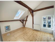 Dachgeschossraum mit Holzbalken und schrägen Wänden, hellem Holzfußboden, einem großen Dachfenster sowie einem seitlichen Fenster mit Sprossen. Ein weißer Heizkörper ist unter dem seitlichen Fenster angebracht.