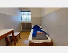 Kleiner, einfacher Gefängnisraum mit einer schmalen Liege auf der rechten Seite, bedeckt mit einer gefalteten Decke und einem blauen Overall. Links steht ein Holzschreibtisch mit einem kleinen Hocker, und das Fenster ist mit Glasbausteinen versehen.