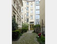 Hinterhof eines Wohngebäudes mit Kopfsteinpflaster, umgeben von historischen Fassaden, bewachsenen Wänden und kleinen Sträuchern. Im Hintergrund befindet sich eine gelbe Eingangstür mit großen Fenstern.