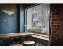 Ein kleines Café mit rustikaler Inneneinrichtung, darunter eine grobe Ziegelwand, ein Holztisch mit Hockern und eine Tischlampe. Durch ein großes Fenster ist eine städtische Straßenszene mit alten Gebäuden, einem Fahrrad und einem Straßenschild zu sehen.