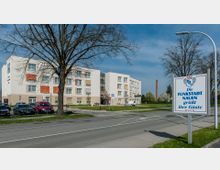 Mehrstöckiges modernes Gebäude mit weißer Fassade und farbigen Fensterläden, umgeben von einer Grünfläche mit Bäumen. Im Vordergrund steht ein Schild mit der Aufschrift „Die Funkstadt Nauen grüßt ihre Gäste“ neben einer Straße und einem Gehweg.