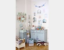 Kinderzimmer mit hellem Holzfußboden und pastellfarbenen Möbeln, darunter eine blaue Kommode und ein kleiner Nachttisch. Spielzeug ist auf Regalen, in einer kleinen Spielzeugkiste und einem Bettenmodell arrangiert, während Bilder und bunte Girlanden die Wände schmücken.