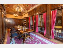 Eleganter Konferenzraum mit Holzwänden, verzierten Deckenmalereien, einem großen Holztisch mit gepolsterten Stühlen und roten Vorhängen an großen Fenstern. Im Raum sind Bücherregale und Kronleuchter, die eine historische Atmosphäre erzeugen.