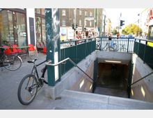 Eingang zu einer U-Bahn-Station mit Treppen, flankiert von Geländern, in einer belebten Stadtumgebung. Im Hintergrund sind Straßencafés, Fahrräder und Geschäfte entlang einer Kreuzung zu sehen.