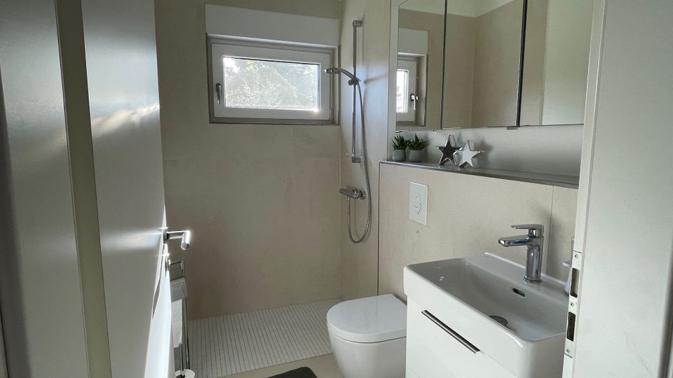 Ein modernes, helles Badezimmer mit beigen Wänden und einem kleinen Fenster. Zu sehen sind ein wandmontiertes WC, ein Waschbecken mit Schubladen darunter, ein Spiegel mit integriertem Stauraum und eine offene Dusche mit einer Handbrause.