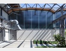 Industriehalle mit modernisierten Elementen; im Vordergrund Betonboden, ein bepflanztes Beet und ein Lüftungsschacht, im Hintergrund eine breite Fassade aus Glas mit vertikalen Streben und einer Stahlträgerkonstruktion unter einem geneigten Dach.