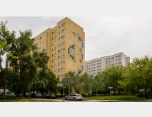 Ein gelbes Hochhaus mit geometrischem Wandmuster aus bunten Fliesen steht in einer grünen Gegend mit Bäumen und parkenden Autos. Im Hintergrund sind weitere Wohngebäude in hellen Farben zu sehen.