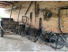 Unterstand mit einer Backsteinwand, an der alte landwirtschaftliche Werkzeuge, Sägeblätter und Ketten hängen. Vor der Wand stehen zwei schwarze Fahrräder sowie ein alter Holzwagen mit Metallrädern.