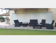 Überdachte Terrasse mit einer Sitzgruppe aus schwarzem Flechtmaterial, bestehend aus Sesseln, Sofas und Couchtischen auf grauen Steinfliesen. Im Hintergrund befindet sich eine Hauswand mit geschlossenen weißen Rollläden, und links ist ein angrenzender Bereich mit zusätzlichen Gartenmöbeln zu sehen.