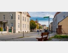 Straßenszene in einem Wohngebiet mit mehrstöckigen Häusern im klassischen Baustil, Gehwegen, parkenden Autos und einer Holzbank im Vordergrund. Ein Schild mit der Aufschrift „Mitfahrbank“ ist neben der Bank angebracht.