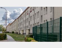 Langes Wohngebäude in schlichter Bauweise mit mehreren Etagen und identischen Fenstern, davor ein Gehweg, Rasenflächen und vereinzelt Fahrräder. Rechts ein hoher grüner Metallzaun, links Straßenlaternen und geparkte Autos, im Hintergrund Bäume und Wolken.